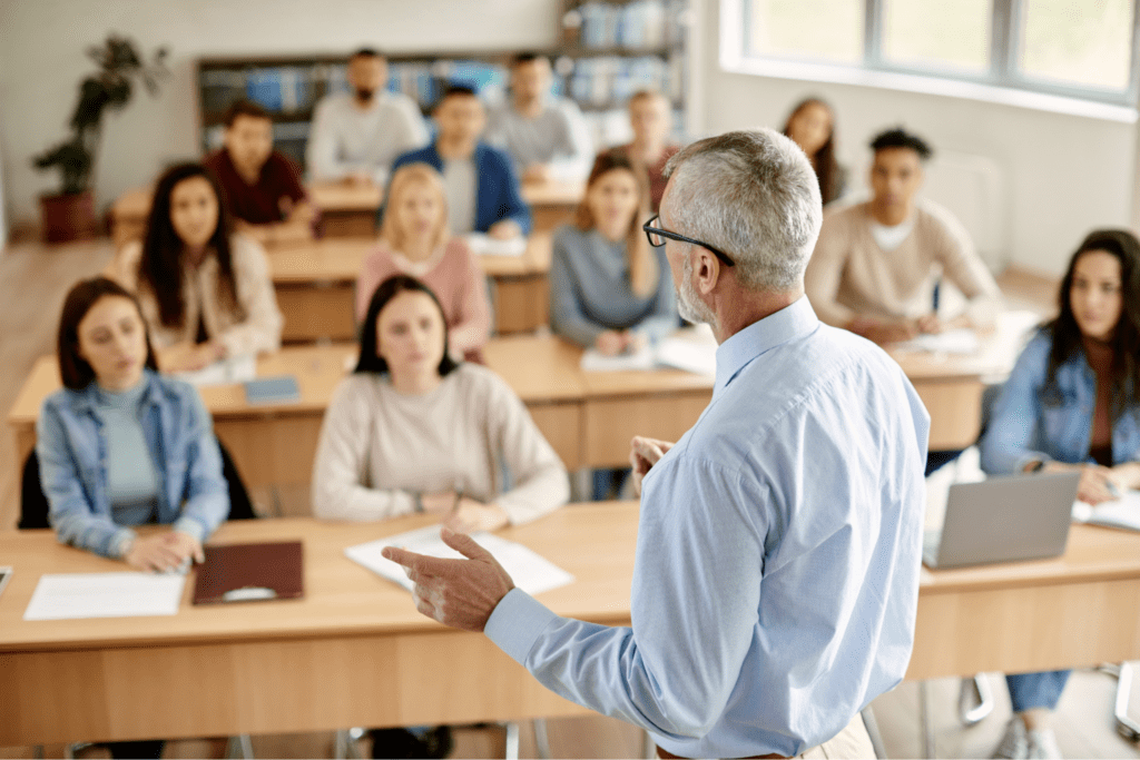 Professor with light skin, grey hair, and glasses, giving lecture to students in bright classroom
